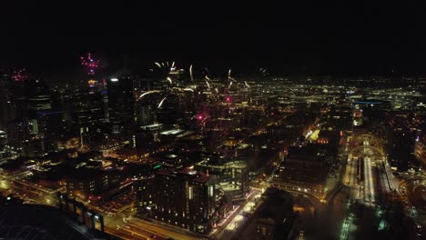 Vista-Aérea-De-Drones-Volando-Hacia-Atrás-A-Través-De-Un-Espectáculo-De-Fuegos-Artificiales-Explosivos-Sobre-El-Horizonte-Del-Centro-De-Denver-Por-La-Noche