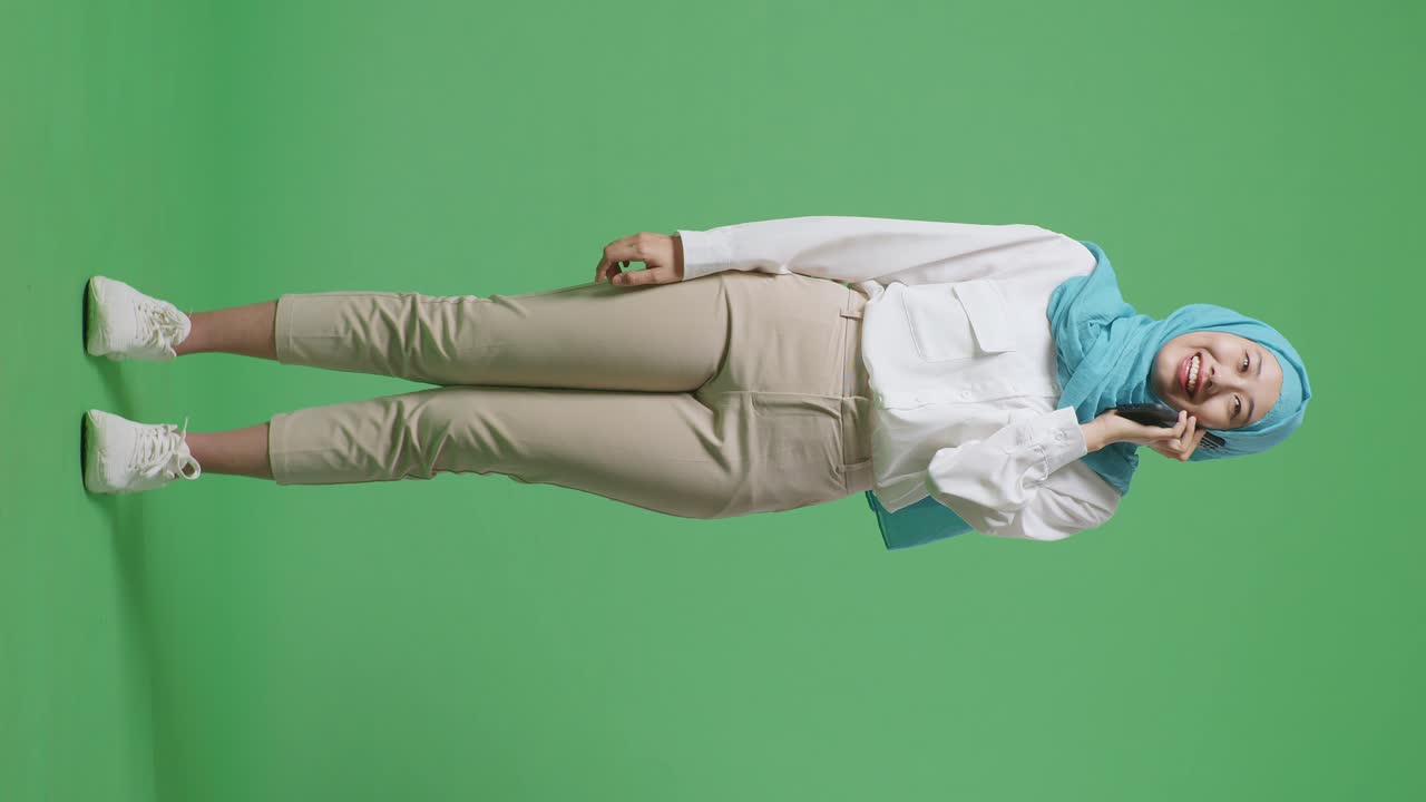 녹색 스크린 배경 스튜디오에 서서 스마트폰으로 이야기하고 미소 짓는 아시아 무슬림 여성의 몸이 가득 차 있습니다.