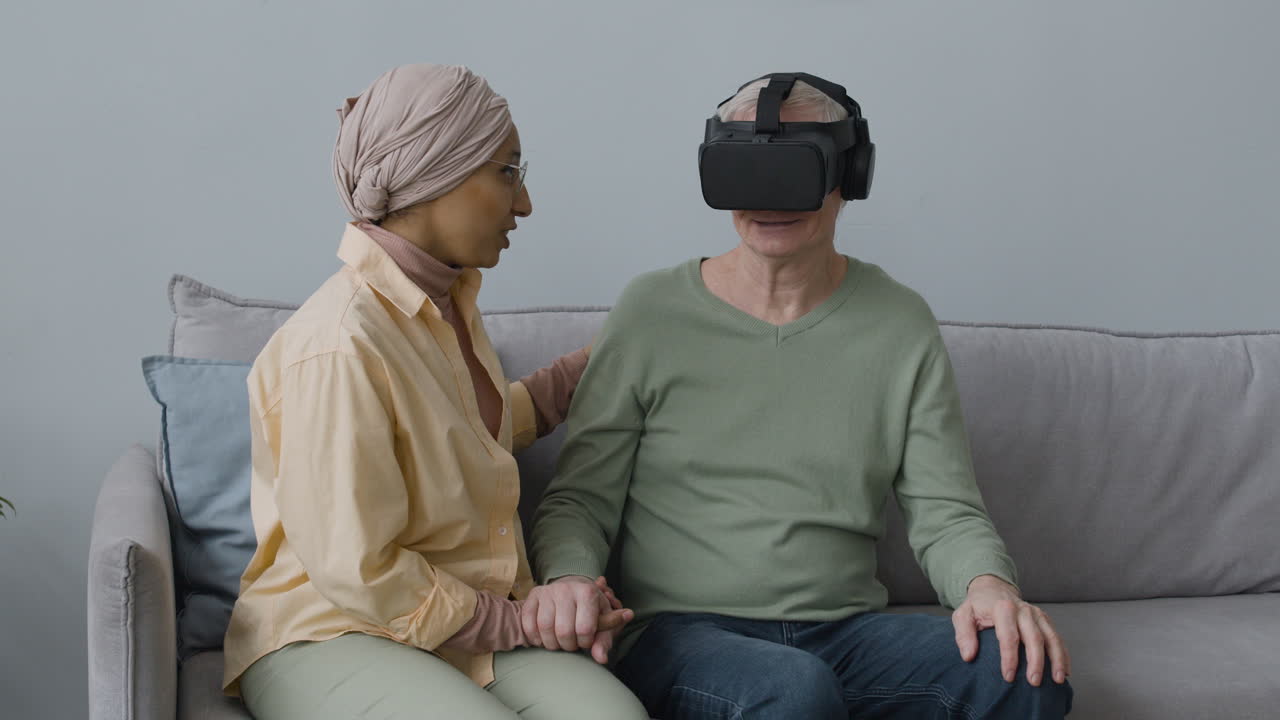 mujer árabe de mediana edad ayudando a un anciano con gafas de realidad virtual mientras se sientan juntos en un sofá en casa