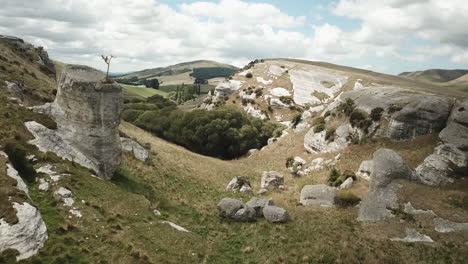 forward drone shot of rocky hills covered in grass, in new zealand, with some meadows and trees, on a cloudy day