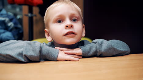portrait of a little boy sitting at the table in the room and looking at camera. curious child with blue eyes talking funny and making different emotions on his small face.