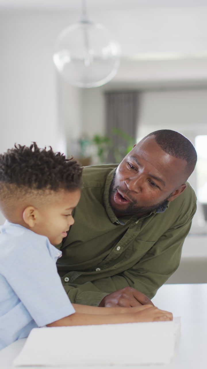 Vertical video of african american man and his son sitting at table and reading braille