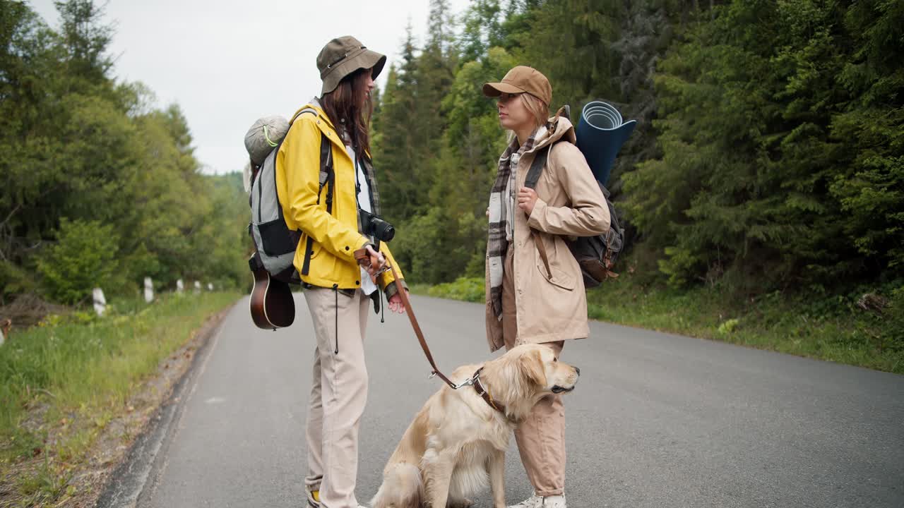 2 chicas en una caminata. una chica morena con una chaqueta amarilla sostiene a su perro de color claro en una correa mientras charla con su amiga cerca de la carretera en el medio del bosque