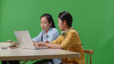asian woman student teaching her friend on a laptop while sitting on a table in the green screen background classroom