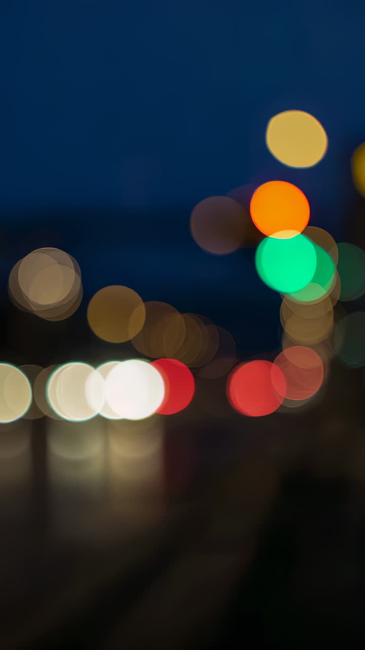 Vertical video: Passing vehicles causing headlights creating bokeh with traffic lights on wet road