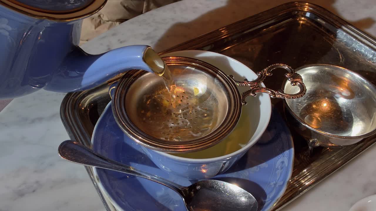Dettagliata immagine dall'alto di tè ambrato che scorre da una teiera in ceramica blu in una tazza attraverso un colino d'argento decorato su un elegante vassoio in un caffè