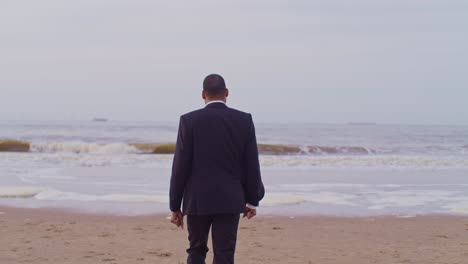 Un-Hombre-Modelo-Latino-Negro-Con-Traje-Camina-Por-La-Playa-Con-Gafas-De-Sol-En-Los-Países-Bajos,-La-Haya,-Vista-Desde-Atrás