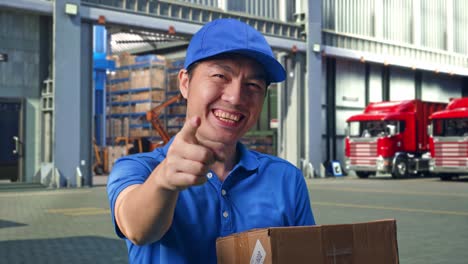 close up of asian male courier in blue uniform smiling and making honest gesture while delivering a carton, outside of logistics distributions warehouse