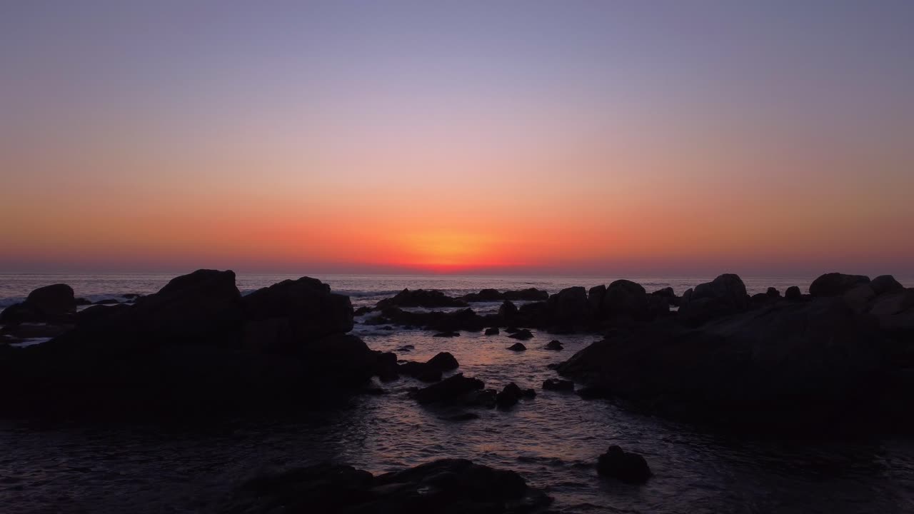 Stunning drone footage flying steadily from the rocky shoreline to the open sea, centered on the vibrant setting sun, capturing the serene beauty of a colorful ocean sunset.