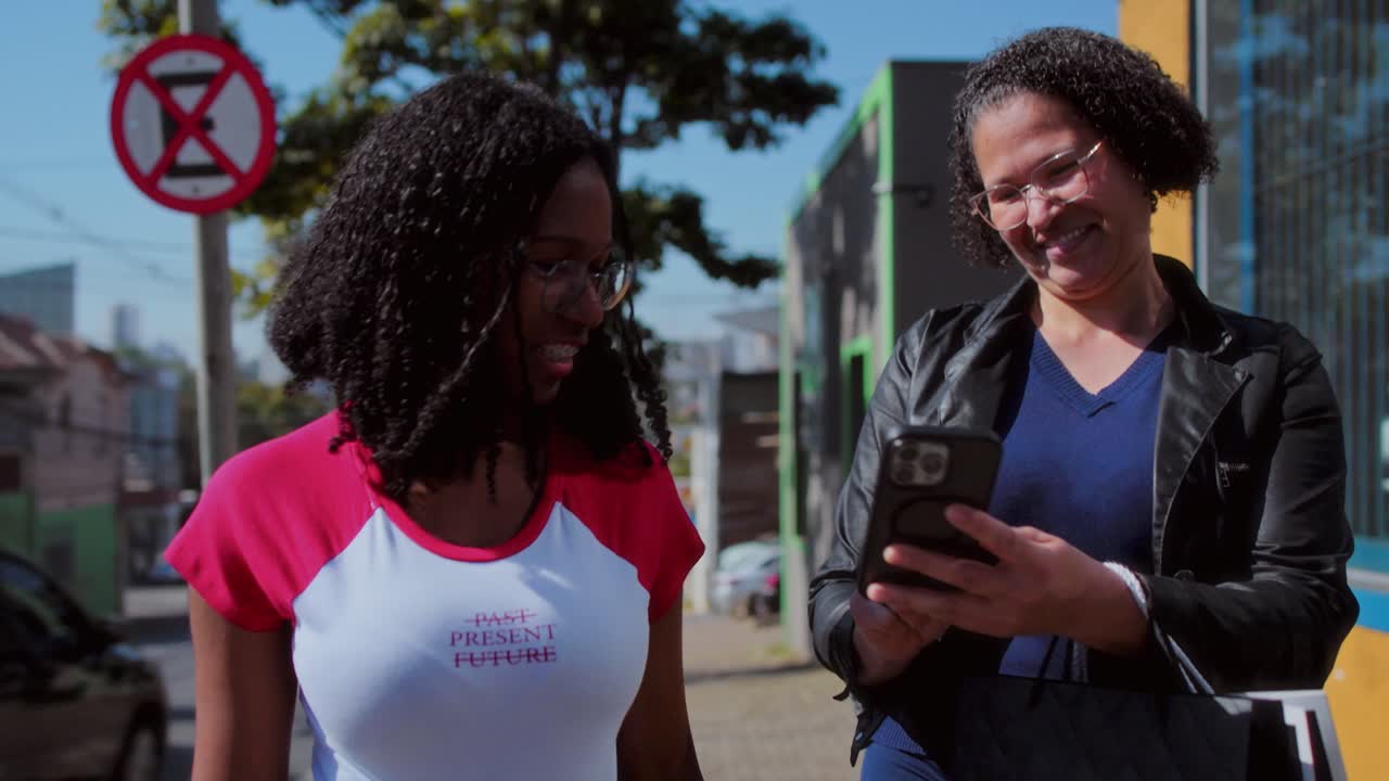 Two women smiling and looking at a smartphone while walking outdoors in the city