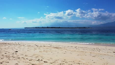 Bali,-Indonesia,-paradise-sandy-beach,-seascape-with-small-tropical-island-and-boats-sailing-in-the-distance,-bright-blue-sky-with-fluffy-white-clouds