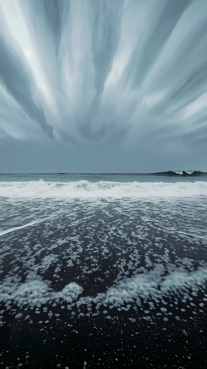 Vertical video: Rolling waves cresting, wind-driven, spilling foam onto pebbly sand, outcrop right