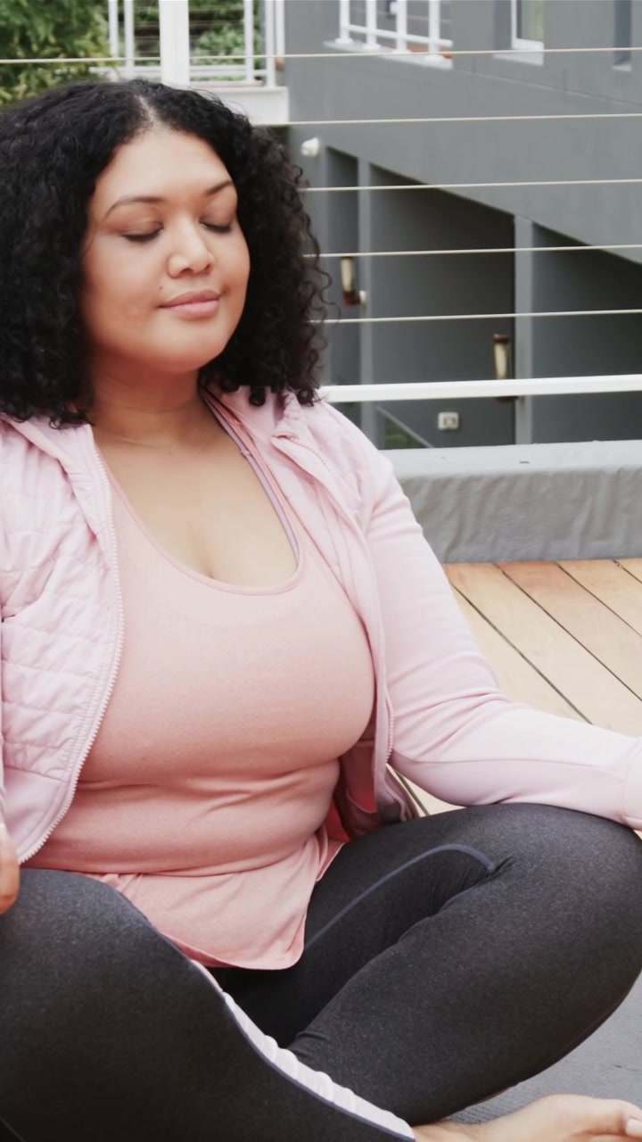 Video verticale: Donna che inspira, seduta a meditare, respirare e calmarsi sul tetto, con giacca rosa e cuscino.