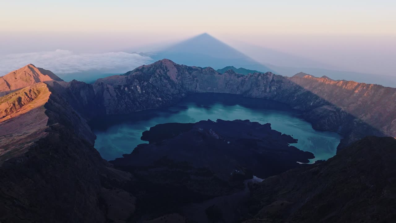 Vista panoramica della caldera vulcanica e del lago craterico del Monte Rinjani all'alba o al tramonto, con un'ombra di vetta prominente.