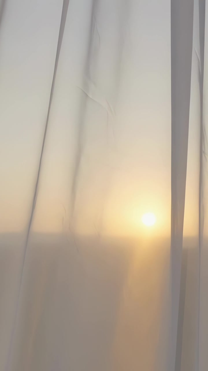 Vertical video: Moving low sun at horizon, sheer curtains swaying at window revealing warm sun disk