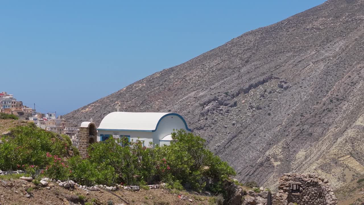 Aerial drone orbit captures a whitewashed chapel and reveals the traditional colorful village of Olympos in Karpathos, surrounded by dramatic mountain landscape