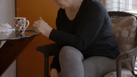 senior woman sits on a comfortable chair, in a cozy kitchen nook. her hemiplegic right hand falls off her lap and to the side, as she reaches for her reading glasses. camera tilts up to her face.