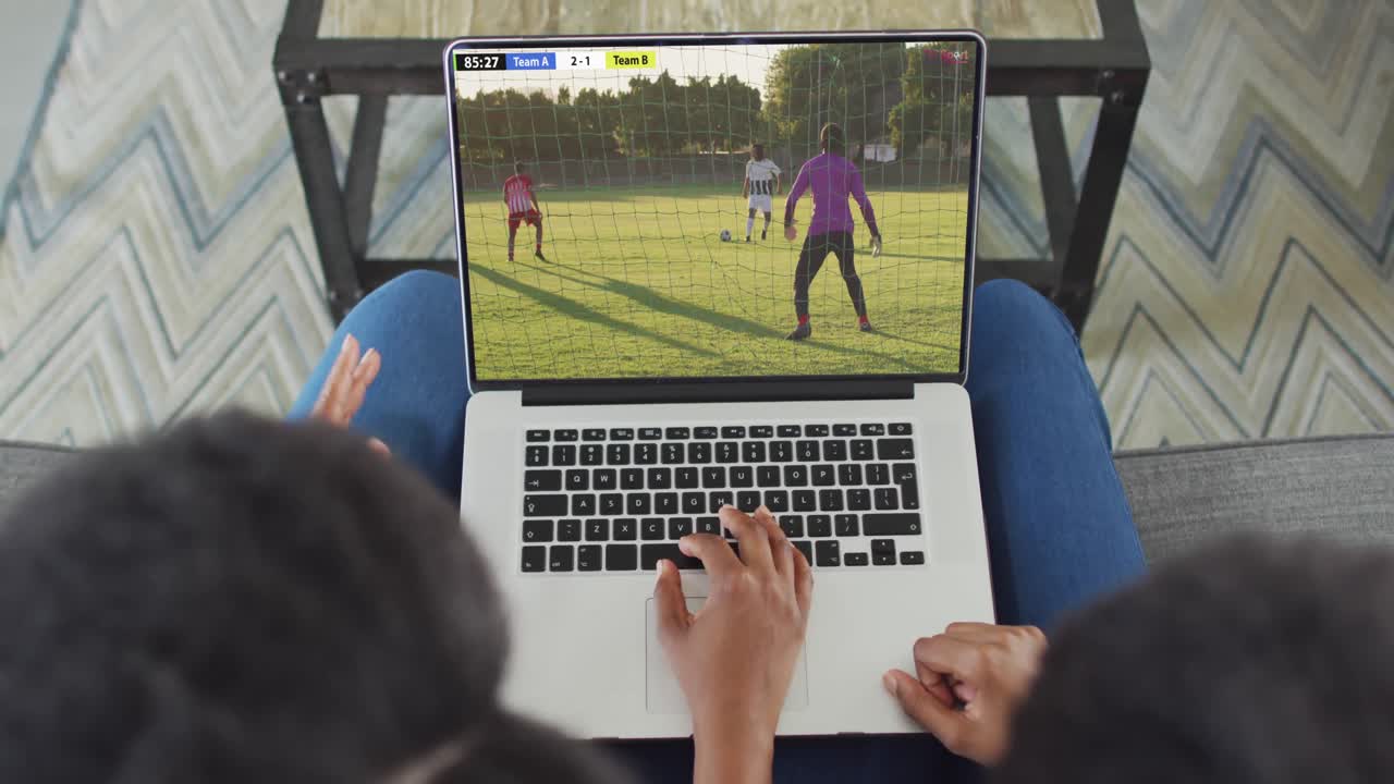 video de dos personas afroamericanas sentadas en el sofá y viendo un partido de fútbol en una computadora portátil