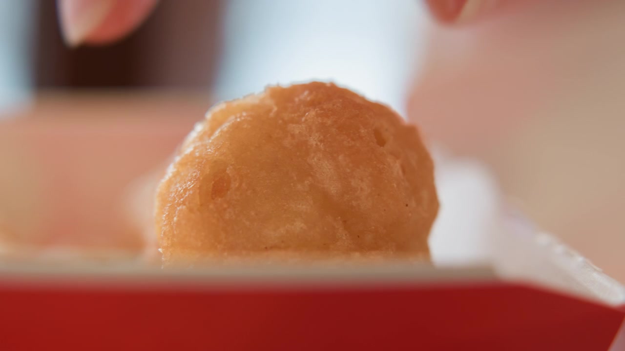 Una mano solleva una singola polpetta di cibo condita dalla fodera di carta bianca all'interno di un contenitore da asporto rosso, mostrando glassa lucida, consistenza cremosa, luce interna soffusa, sottile sfocatura di movimento nel punto di contatto e fresco