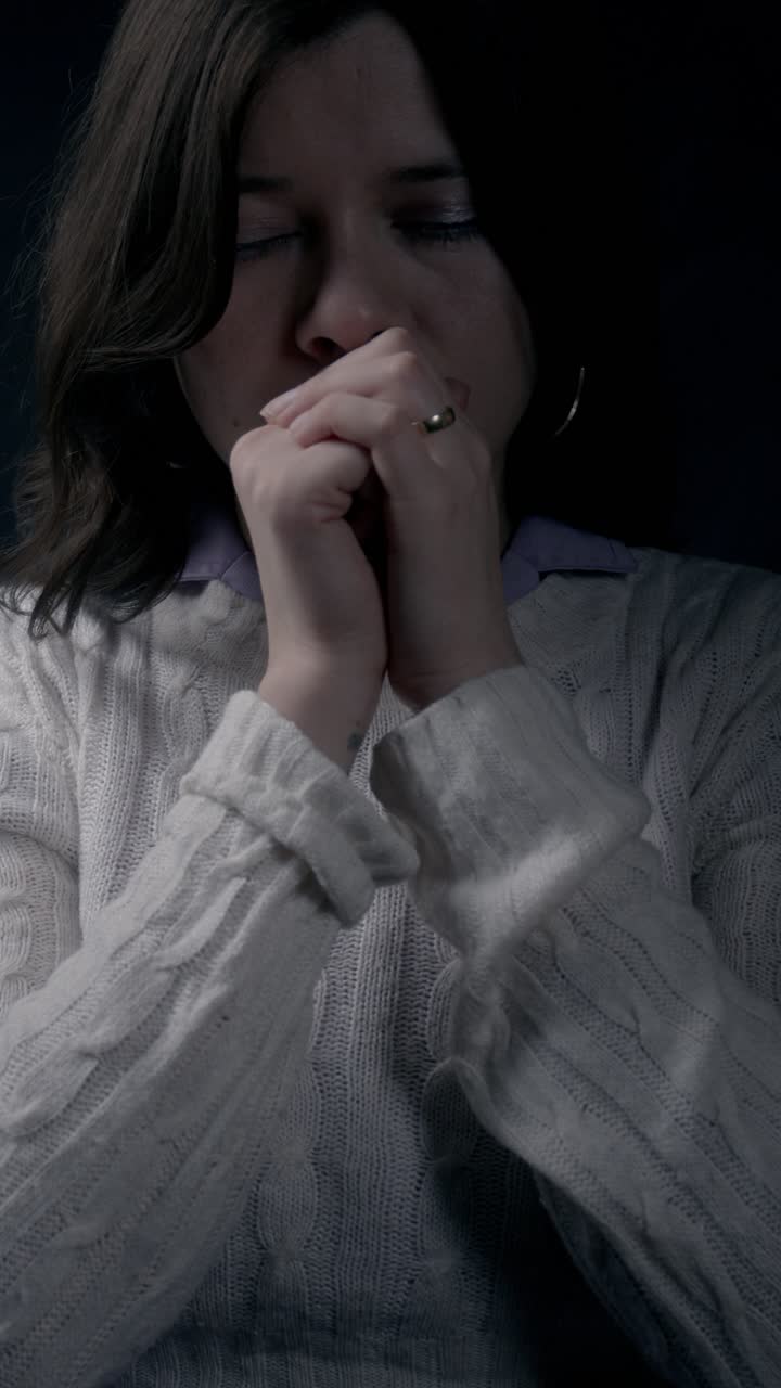 Vídeo vertical íntimo e em close-up de uma mulher latina em oração, com as mãos juntas, transmitindo uma emoção profunda. Pouca luz, capturando a atmosfera serena e contemplativa perfeitamente para stories no celular