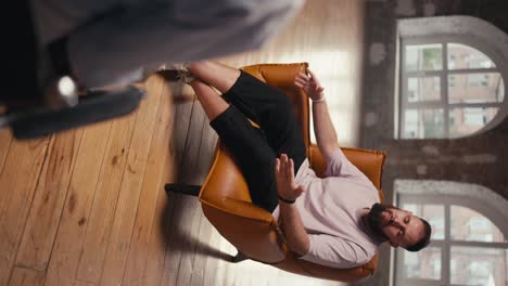 Vertical video of a man with a beard in a light pink T-shirt and black shorts sitting on a light brown table talking to a psychologist about his problem during therapy