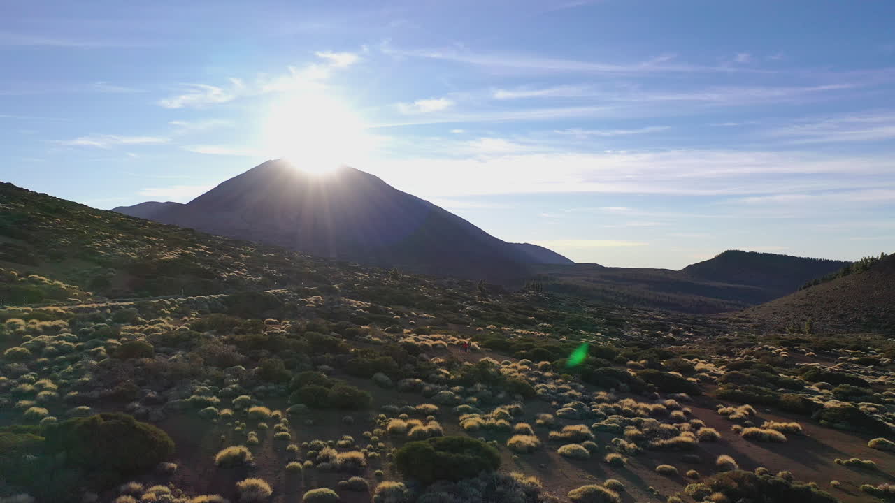 Three backpackers standing in the bushes on the hillside below the Pico de Teide mountain on Canary Islands and watching the sun setting behind the mountain peak. Hikers watching a mountain sunset 4K.