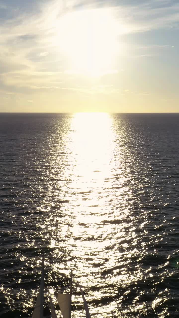 A sailboat sails into the distance with its silhouette framed against a glowing horizon, where sunlight scatters across the rippling ocean in dazzling reflections
