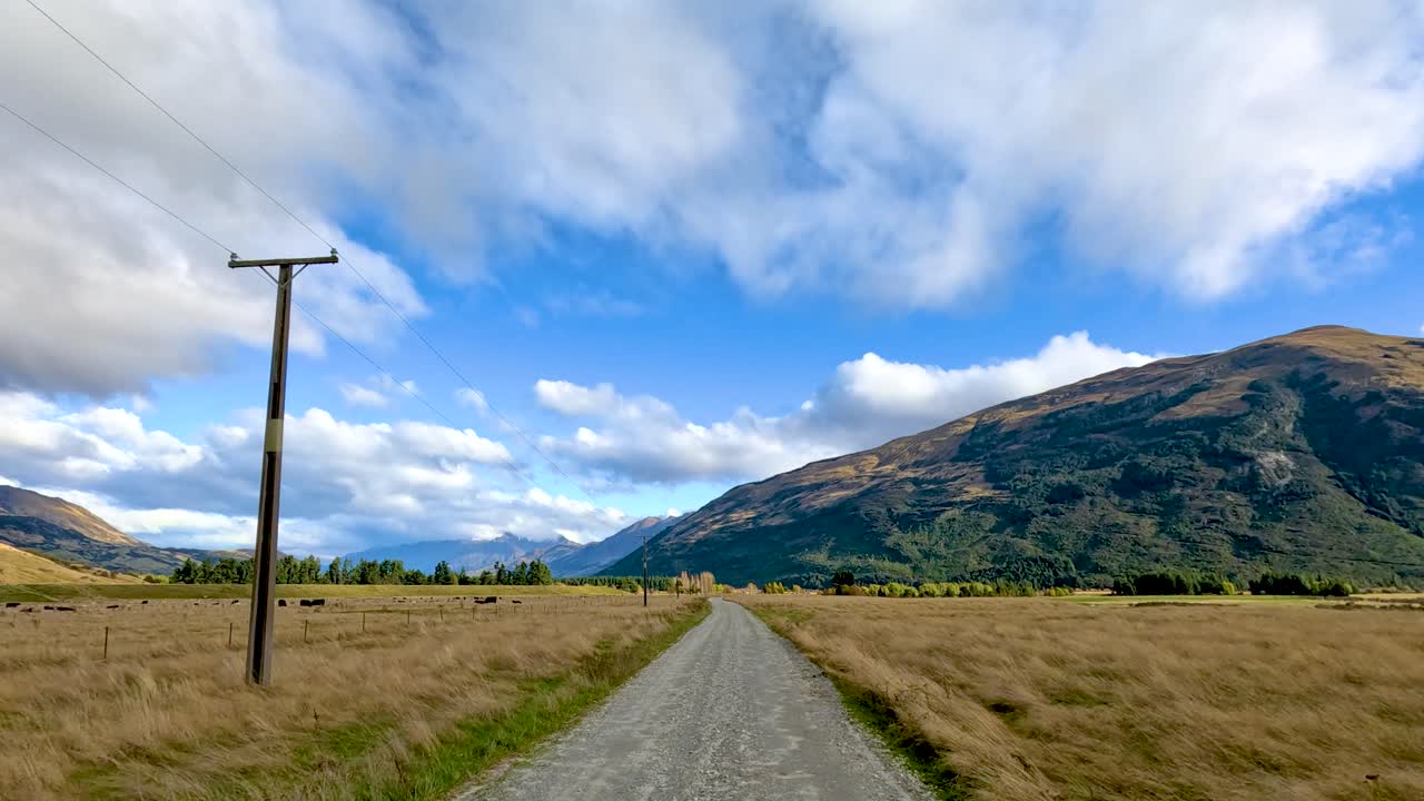 Vehicle travels straight on rural gravel road, passing fields and mountain under bright daylight sky