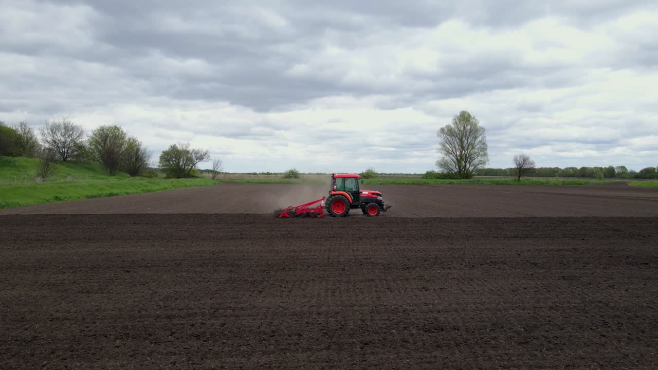 una toma aérea de un tractor arando un pequeño campo con polvo soplando en el campo de una pequeña ciudad.