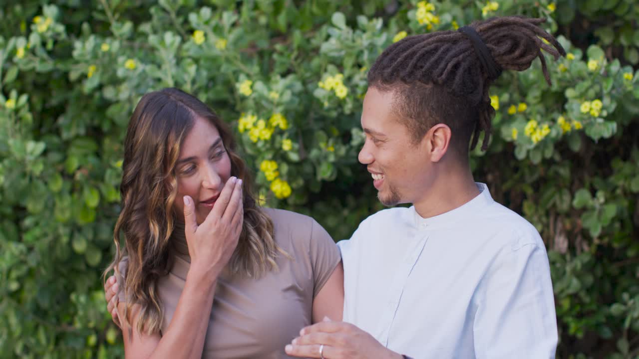 Diverse couple by flowered hedge, man leaning in flirting, woman laughing, wristwatch visible