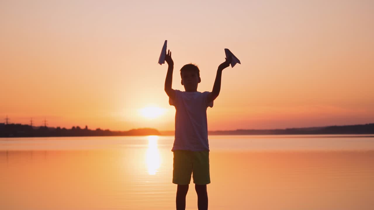 Boy standing with paper planes near the river. Child playing with origami planes in slow motion on the evening river background.
