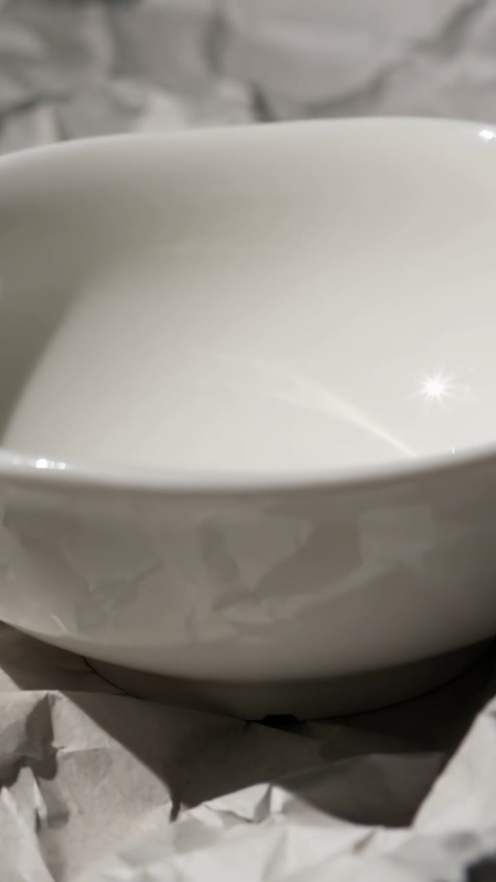 Vertical video: Moving camera revealing white ceramic bowl on crumpled paper, showing glossy rim