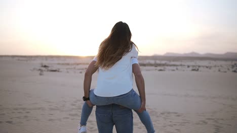 happy and cute adorable adult couple in white shirts and jeans man with woman girlfriend on piggy back running by desert crazy in love, emotions and relationship. whirling around. mild sunset on background