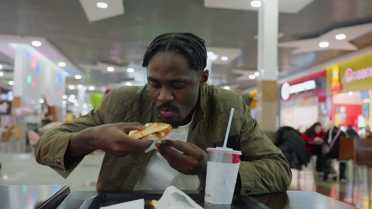 Young man savoring burger while seated at food court table with drink cup and tray, wearing green jacket, casual dining atmosphere, blurred background with people and lights