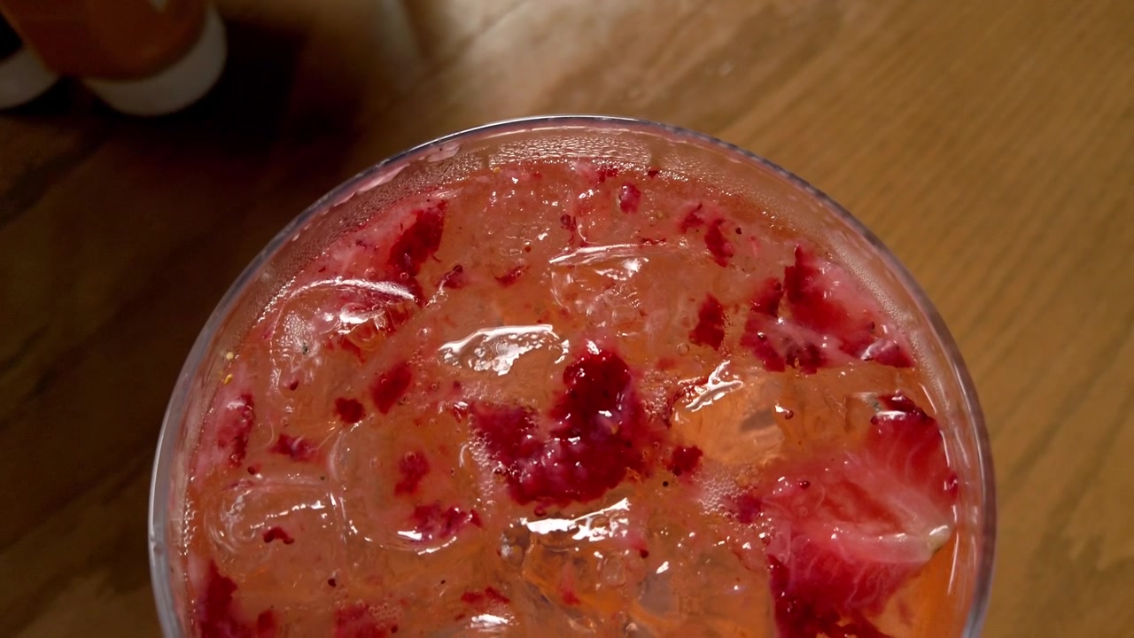 Vista superior aproximada de bebida de fruta rosa com gelo picado em copo sobre mesa de madeira. Pedaços de fruta vermelha e polpa são visíveis por toda a bebida gelada