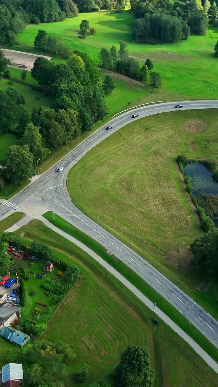 Aerial landscape revealing winding road near Talsi, Latvia, with vehicles traversing green countryside, residential areas, and tranquil water body, drone shot