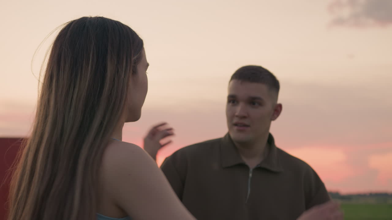 young couple enjoys tender moment after hot air balloon landing as woman affectionately reaches toward man during calm evening in open field with pastel sky and soft sunset light in background