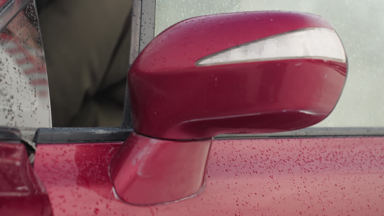 Close up of red car side mirror and door with water droplets on surface during winter as boy steps out of vehicle, with soft blur background and cold seasonal atmosphere reflected on glass