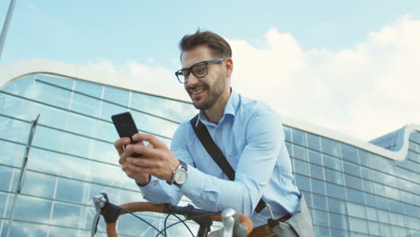 Camera-rolling-round-a-handsome-man-wearing-glasses-and-shirt-while-texting-on-the-smartphone-device-and-sitting-on-his-bike-in-the-city-center