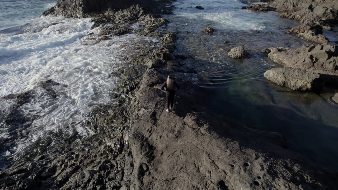 Ripresa aerea in orbita di una giovane donna che si trova tra le piscine naturali e dove si possono vedere le onde del mare.