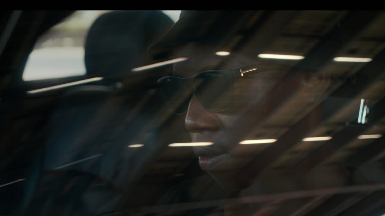 차에서 선글라스를 입은 흑인 남자가 누군가를 스파이하는 것처럼 측면 시야 거울을 보고 있습니다.