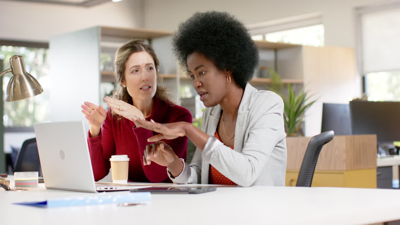 Diverse female creative colleagues in discussion using laptop in office, slow motion