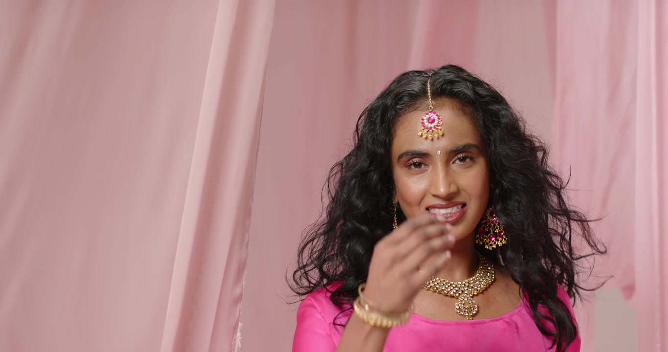 Responding to cue, Indian woman peeking from studio pink drapes, adjusting tikka, smiling at camera