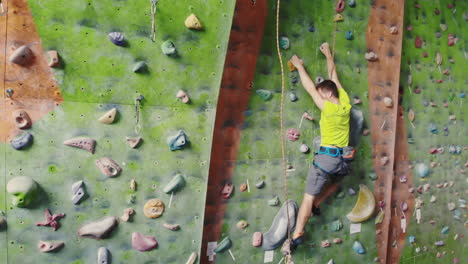 Male-climber-climbs-the-cliff-wall-in-the-room-reaching-and-gripping-hold.-A-professional-concentrated-climber-moves-up-the-wall-the-camera-rises-and-lays-down-his-movements.-Boom-and-truck-follow-camera.