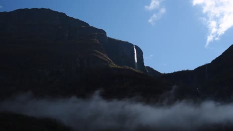 Blick-Auf-Eine-Dunkle-Bergkette-Mit-Einem-Sonnenbeschienenen-Wasserfall-Und-Wolken
