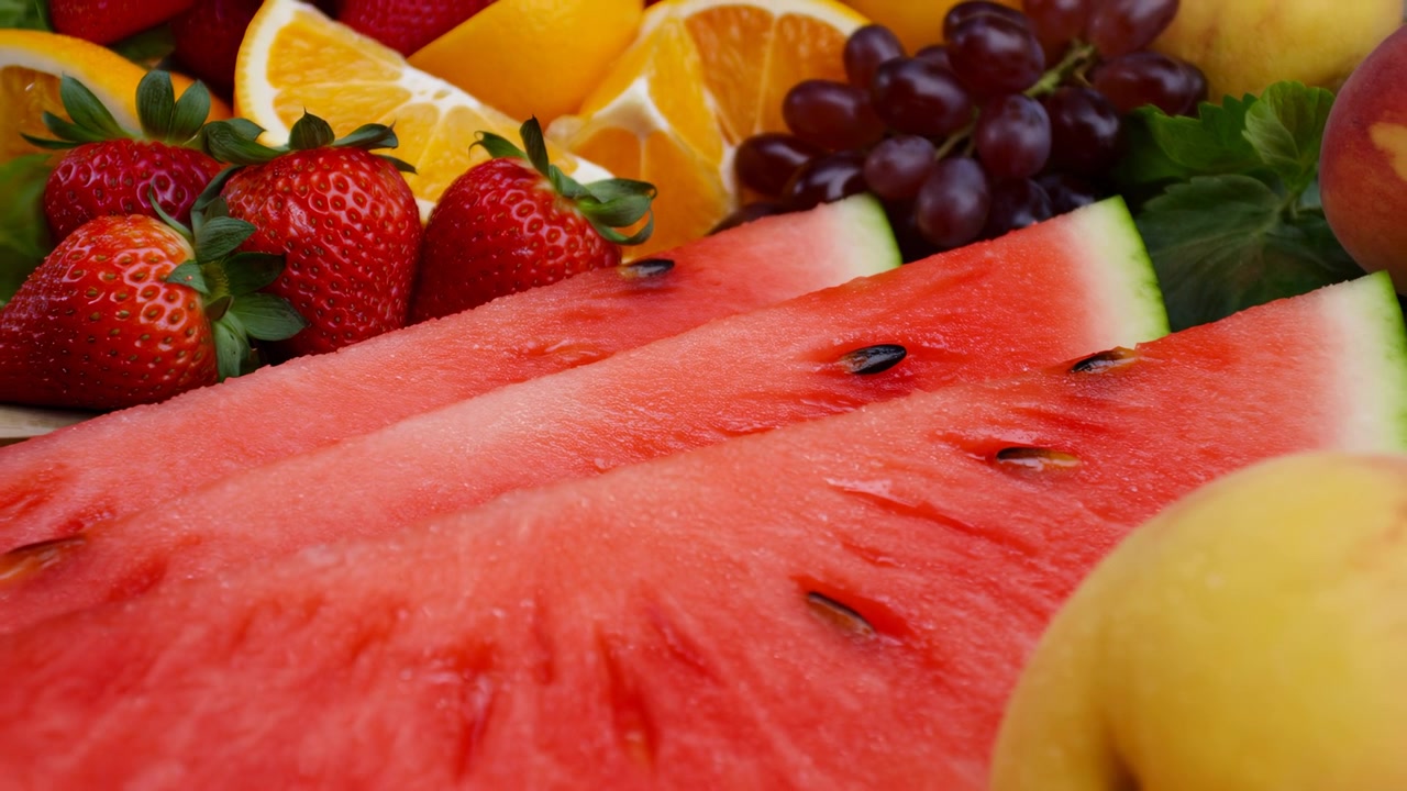 Slow pullback widens close view of juicy watermelon with visible seeds, bringing strawberries, citrus wedges, grape clusters, and surrounding leafy greens into fuller colorful composition.
