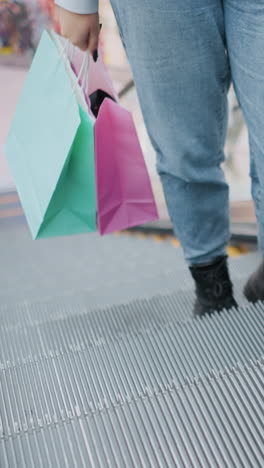 vista de la pierna de una persona en vaqueros y botas negras sosteniendo bolsas de compras, balanceándose suavemente mientras sube una escalera mecánica en un bullicioso centro comercial con luces brillantes y diseño interior moderno