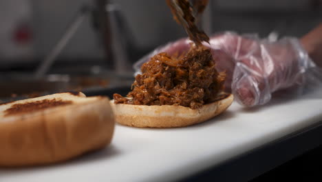 cook uses tongs to place chopped beef on toasted bun, barbecue sandwich, close up slow motion 4k