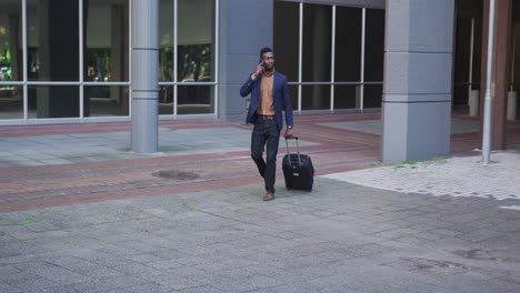 african american businessman using smartphone walking with suitcase in street