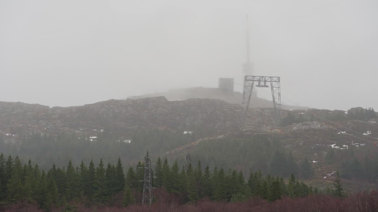 Ulriken mountain in Bergen Norway, large mast on top and a cable car ascending through fog and rain blowing sideways